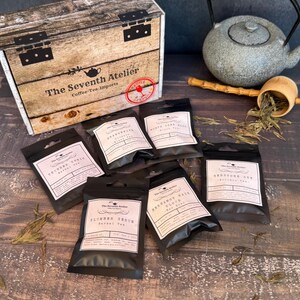 May include: A wooden box with the text "The Seventh Atelier Coffee-Tea Imports" sits next to several black tea packets. A gray teapot, bamboo scoop, and loose tea leaves are also present, suggesting a tea-themed product.