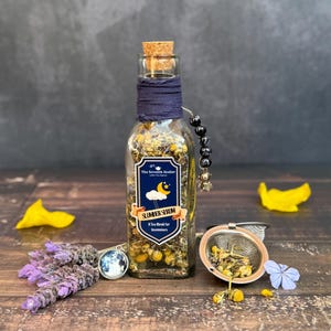 May include: A clear glass bottle filled with dried herbs and a cork stopper. The bottle is labeled "Slumber Serum" and wrapped with navy blue fabric. A small tea infuser, lavender sprig, and a charm bracelet are also present.