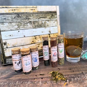 May include: A set of six small glass bottles with cork tops filled with loose leaf tea. The bottles are labelled with names such as "Evening Star" and "King's Field". The tea is displayed on a wooden surface with a mug of tea and a tea infuser.