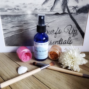 Puede incluir: Una botella azul de spray fijador de maquillaje con el texto "Surf's Up Essentials" y "Make Up Setting Spray" en la etiqueta. La botella está al lado de un tarro rosa y un tarro dorado de polvo de maquillaje. Hay dos pinceles de maquillaje y una piedra blanca sobre una superficie de madera.
