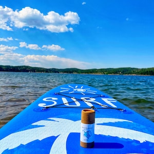 May include: A brown cardboard tube with a white label that says "Natural Sunscreen" and has a picture of a sea turtle. The tube is sitting on a blue paddleboard with the word "SURF" written on it. The paddleboard is floating on a lake with a blue sky and white clouds in the background.