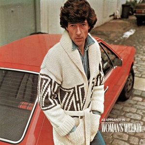 以下が含まれることがあります： 白地に茶色と白の幾何学模様が施された白いニットカーディガンを着た男性。カーディガンはショールカラーでベルト付き。男性は赤い車の横に立っている。画像の右下には「AS APPEARED IN WOMAN'S WEEKLY」というテキストが表示されている。