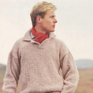 May include: A beige knitted jumper with a collared neckline and a single button. The person is wearing a red scarf and blue jeans. A plaid hat is held in the hand.