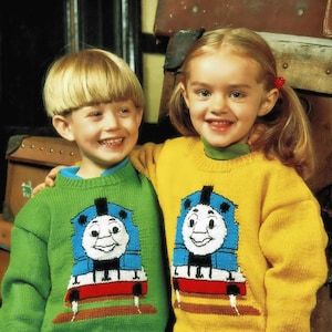 May include: Two children wearing knitted sweaters with Thomas the Tank Engine designs. The boy is wearing a green sweater with a blue and white Thomas design. The girl is wearing a yellow sweater with a blue and white Thomas design.