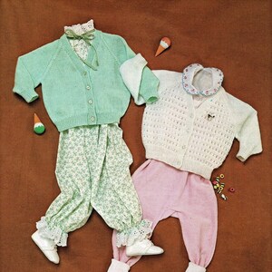 May include: Two knitted baby cardigans, one green and one white, with matching pants. The green cardigan has a white collar and a green ribbon. The white cardigan has a white collar with a blue trim. Both cardigans have buttons down the front.