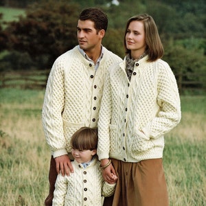 May include: A family of three wearing cream-coloured cable knit cardigans. The adults are holding hands with a young child. The child is wearing a cream-coloured cable knit cardigan and brown trousers.