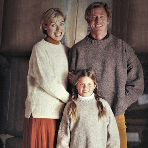 Pode incluir: Uma família de três pessoas vestindo suéteres de tricô. A mulher está vestindo um suéter branco com uma saia longa laranja. O homem está vestindo um suéter marrom com calças de veludo cotelê marrom. A criança está vestindo um suéter bege claro com jeans azul.