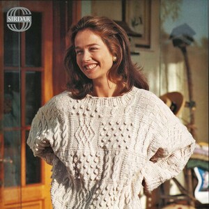 Puede incluir: Suéter de punto color crema con cuello redondo y mangas cortas. El suéter presenta varios patrones de trenzas y puntos de fantasía. El logotipo de Sirdar es visible. El suéter se combina con una falda estampada.