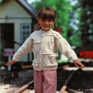 May include: A young girl wearing a white knitted cardigan with a floral design and red and white checkered pants. She is standing on a set of railroad tracks.