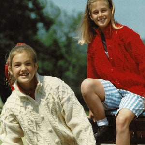 May include: Two young girls wearing sweaters. One girl is wearing a white cable knit sweater and a floral patterned skirt. The other girl is wearing a red cardigan and blue and white striped shorts. Both girls are sitting on a bale of hay.