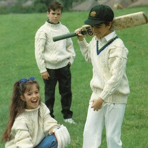 May include: Three children are playing cricket on a grassy field. A girl is kneeling down next to a gray and green duffel bag. A boy is holding a cricket bat over his shoulder. Another boy is standing behind the girl, looking at the camera. All three children are wearing white sweaters.