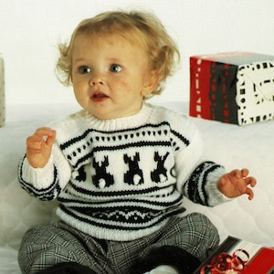 Peut inclure: Un bébé portant un pull en tricot blanc et noir avec un motif de renne. Le pull a une bordure noire et blanche autour du cou et des poignets. Le bébé est assis sur une surface blanche avec des cadeaux emballés en arrière-plan.