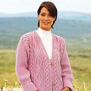 May include: A pink cable knit cardigan with a V-neck, button closure, and pockets. The cardigan is worn by a person standing in a field.