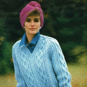 以下が含まれることがあります： Vネックのライトブルーのケーブルニットセーター。青い襟付きシャツと合わせている。モデルはピンクのターバン、ピンクの手袋、チェック柄のパンツを着用。セーターはテクスチャード加工の模様が特徴。