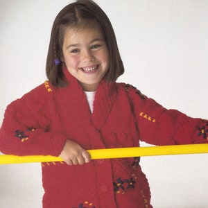 May include: A young girl wearing a red cardigan with embroidered flowers and pink pants. She is holding a yellow bar in front of her.