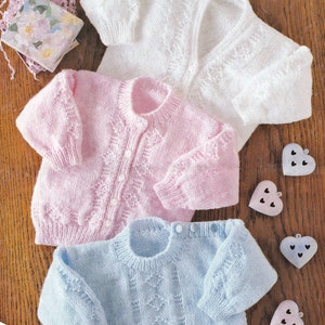 Puede incluir: Tres suéteres de bebé tejidos en blanco, rosa y azul claro, con patrones decorativos. El suéter rosa tiene botones en la parte delantera. El suéter azul claro tiene botones en el hombro. El texto "PREMATURE SIZE INCLUDED" está impreso.
