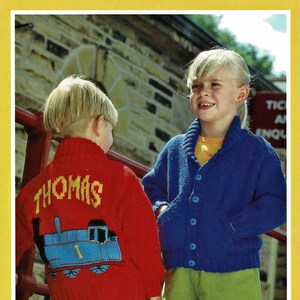Puede incluir: Dos niños con suéteres de punto. El niño lleva un suéter rojo con un diseño de Thomas la locomotora de tanque azul y amarillo y el texto "THOMAS" en la espalda. La niña lleva un suéter azul con cuello amarillo y pantalones verdes.