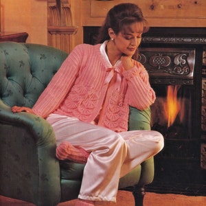 May include: A woman wearing a pink knitted cardigan with a delicate lace pattern and matching pink satin trousers. She is sitting in a green armchair with her feet resting on a rug in front of a fireplace.