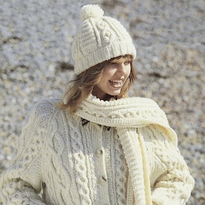 May include: A white knitted cardigan with a cable knit pattern, featuring a matching scarf and a white knitted hat with a pom pom.