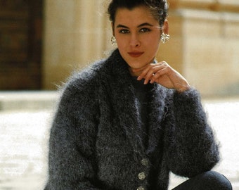 May include: A dark grey fuzzy knit cardigan with a shawl collar and button fastening. The cardigan is being worn by a person with dark hair and is sitting on a stone surface.