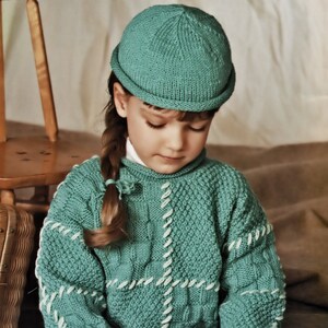 May include: A child wearing a teal knitted hat and sweater with white stitching detail. The sweater has a textured pattern. The child is sitting cross-legged and playing with wooden toy cars and blocks. The background is a neutral color.