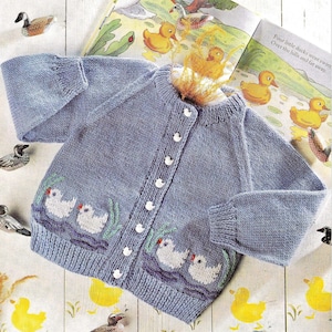Può includere: Un cardigan di lana blu chiaro per neonato con bottoni bianchi e anatre ricamate sul davanti. Il cardigan è steso su una superficie di legno bianco con illustrazioni di anatre gialle.