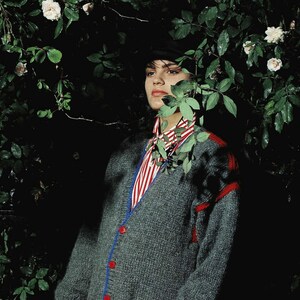 May include: A woman wearing a gray knitted cardigan with red and blue trim, a red and white striped shirt, and a red skirt. She is standing in front of a green leafy background.