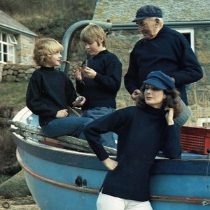 May include: Four people stand on a blue boat. A woman in a black turtleneck sweater and white pants stands on the boat. A man in a blue cap and a black sweater stands behind her. Two children, one in a blue sweater and one in a black sweater, stand in front of the woman.