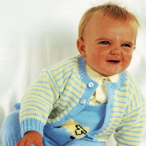 May include: A baby wearing a blue and yellow striped knitted cardigan and matching blue knitted pants with a yellow lamb design. The baby is sitting on a white surface.