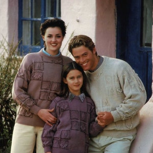 Puede incluir: Tres personas con suéteres de punto a juego con patrones cuadrados y de hojas. Los suéteres son en tonos malva y crema. Las personas están de pie frente a un edificio rosa con una puerta azul.