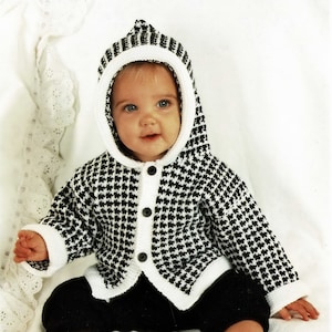 May include: A baby wearing a black and white houndstooth hooded cardigan with white trim and black buttons. The cardigan is knitted and the baby is sitting on a white surface.