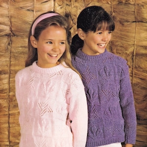 Könnte beinhalten: Zwei Mädchen tragen Strickpullover. Das Mädchen links trägt einen rosa Pullover mit Sternmuster. Das Mädchen rechts trägt einen lila Pullover mit Herzmuster.