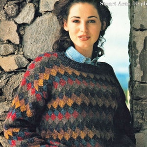 May include: A woman wearing a brown, blue, red and yellow patterned sweater with a chevron design. The sweater is made of a soft, textured yarn. The woman is standing in front of a stone wall. The text "Stuart Aran 10 ply" is visible in the upper right corner of the image.