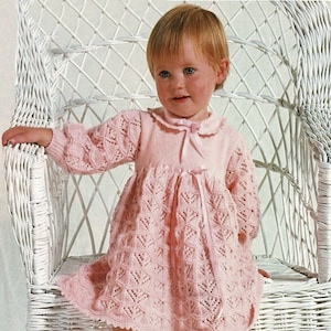 May include: A little girl wearing a pink knitted dress with a lace pattern. She is sitting in a white wicker chair.