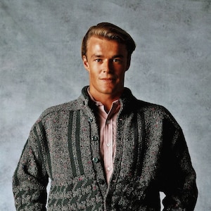 May include: A man wearing a green and black patterned cardigan with a pink button-down shirt underneath. He is wearing dark green pants and has his hands in his pockets.