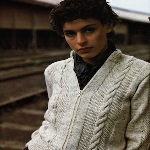 May include: A white cable knit cardigan with a zipper closure. The cardigan has a subtle speckled pattern and a black and white patterned scarf underneath.