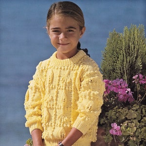 Puede incluir: Una persona joven lleva un suéter de punto amarillo con un patrón texturizado y una falda de estampado blanco y negro. La persona está sentada frente a un arbusto verde con flores rosas.