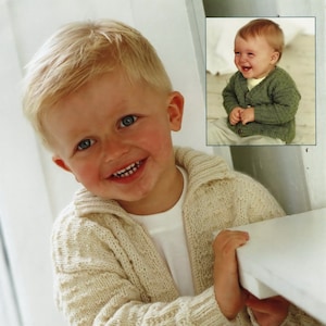 May include: A young boy wearing a cream coloured knitted cardigan with a zip. The cardigan has a cable knit pattern. The boy is smiling and holding onto a white surface.