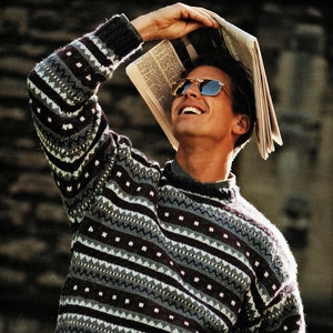 May include: A man wearing a grey and black striped jumper with a white and black pattern. He is wearing sunglasses and holding a newspaper over his head.
