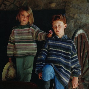May include: Two children wearing striped sweaters stand in front of a wooden wheel. The girl on the left is wearing a pink and green striped sweater and holding a straw hat. The boy on the right is wearing a blue and gray striped sweater and has his hand on a horseshoe.