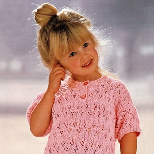 May include: A young girl wearing a pink crocheted sweater with a button fastening and light blue trousers.