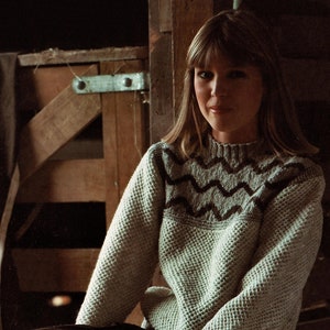 May include: A woman wearing a light gray sweater with a dark gray and white wavy pattern on the neckline. She is sitting with her legs crossed and her hands in her lap.