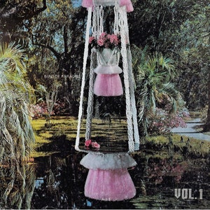 May include: A vintage book cover featuring a white macrame plant hanger with pink tassels and a pink flower pot. The hanger is suspended in front of a green and blue landscape with trees and a pond. The book title is "Classic's Macrame Premiere" and the volume number is "Vol. 1".