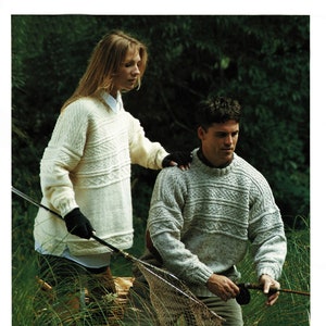 May include: Two people wearing knitted jumpers stand in a grassy area. The person on the left is wearing a white jumper with a cable knit pattern. The person on the right is wearing a grey jumper with a cable knit pattern. They are standing near a fishing net and a fishing rod.