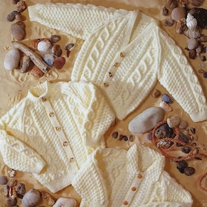 Könnte beinhalten: Drei weiße Strickjacken mit Knöpfen. Die Jacken liegen auf einer sandigen Oberfläche mit kleinen Steinen und Muscheln.