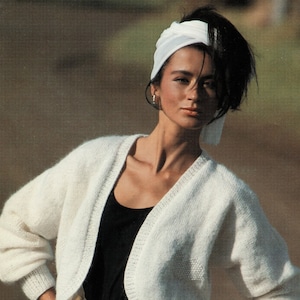 May include: A white cropped cardigan with a V-neckline, worn over a black tank top. The model is wearing a white headband and a gold belt with a large buckle. She is also wearing several gold bracelets on her wrist.