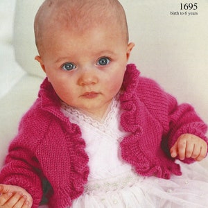 Könnte beinhalten: Ein Baby trägt eine rosa gestrickte Strickjacke mit Rüschenbesatz. Die Strickjacke wird über einem weißen Kleid mit Spitzenbesatz getragen.