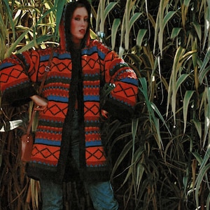 Pode incluir: Uma mulher vestindo um suéter longo com capuz preto estampado em vermelho, azul e verde. Ela está usando jeans azul e botas vermelhas. A mulher está em pé em um campo de plantas verdes altas.