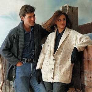 May include: A couple wearing casual winter clothing. The man is wearing a dark gray cardigan over a blue denim shirt and blue jeans. The woman is wearing a white and cream speckled cardigan with a large collar over a black shirt and black pants.