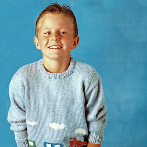 Peut inclure: Un pull en tricot bleu clair avec un motif de trois voitures et de nuages sur le devant. Le pull est porté par un jeune garçon aux cheveux courts bruns. Le garçon sourit et a les mains dans les poches.